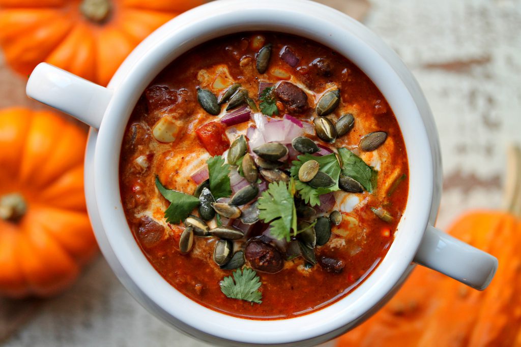 Bowl of soy chorizo and pumpkin chili with toppings of vegan sour cream, red onion, cilantro, and pumpkin seeds on a rustic table setting.