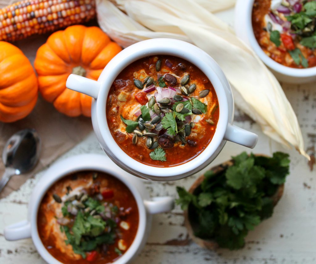 Bowl of soy chorizo and pumpkin chili with toppings of vegan sour cream, red onion, cilantro, and pumpkin seeds on a rustic table setting.