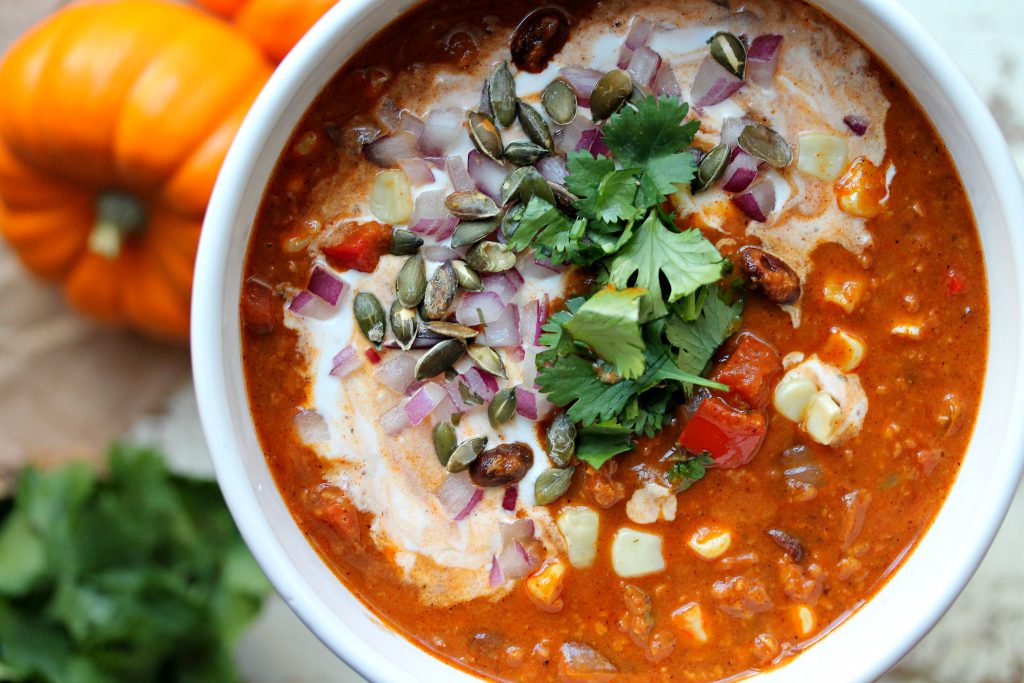 A hearty bowl of vegan soy chorizo and pumpkin chili topped with vegan sour cream, chopped cilantro, red onion, and seasoned pumpkin seeds.