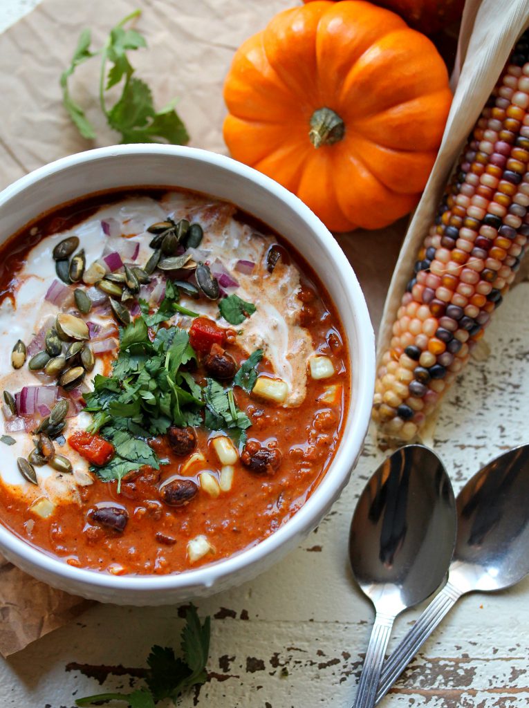 A hearty bowl of vegan soy chorizo and pumpkin chili topped with vegan sour cream, chopped cilantro, red onion, and seasoned pumpkin seeds.