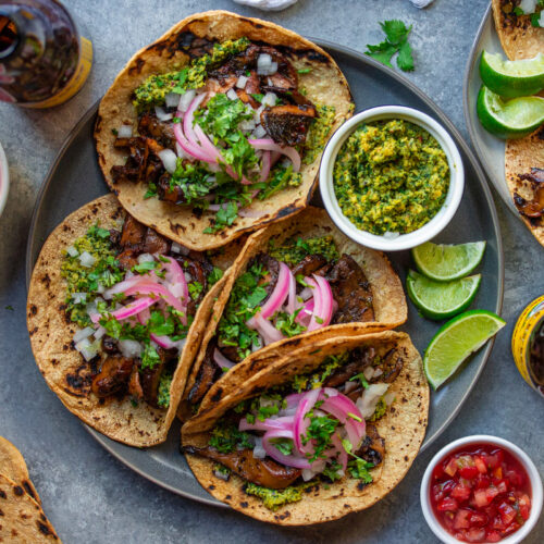 A plate of finished Vegan Asada Mushroom Tacos garnished with fresh lime wedges, cilantro, and hot sauce on the side.