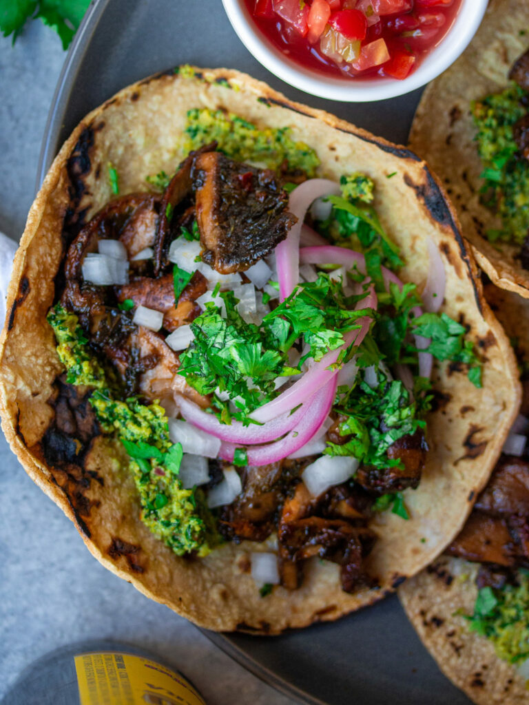 A plate of finished Vegan Asada Mushroom Tacos garnished with fresh lime wedges, cilantro, and hot sauce on the side.