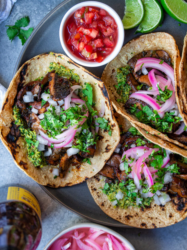 A plate of finished Vegan Asada Mushroom Tacos garnished with fresh lime wedges, cilantro, and hot sauce on the side.