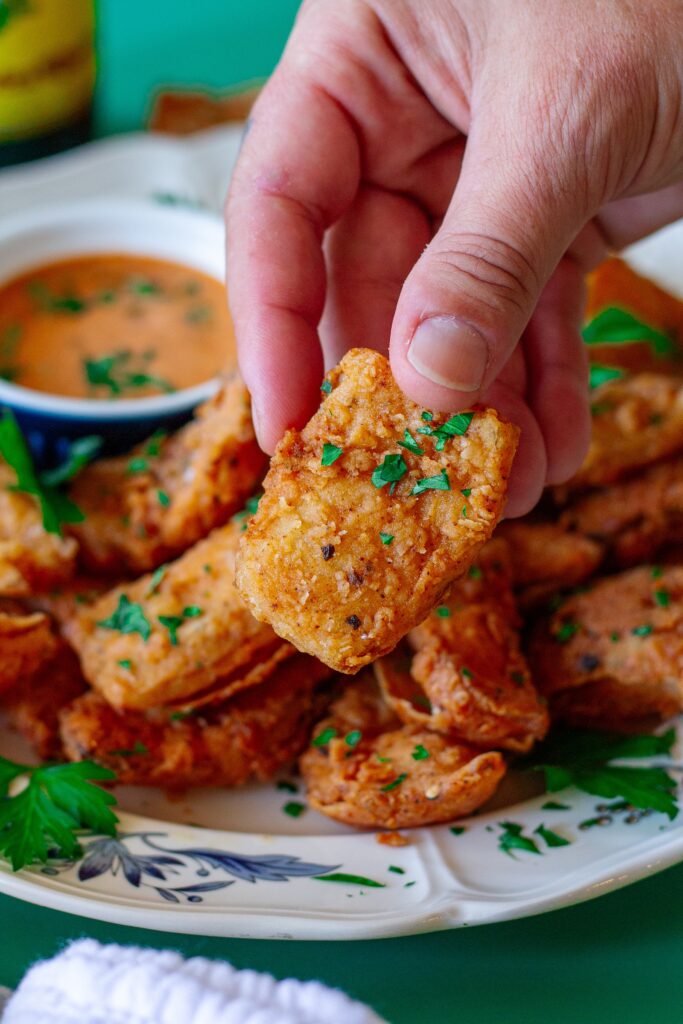 Hand holding a crispy vegan bloomin' onion petal garnished with parsley, ready to be dipped.