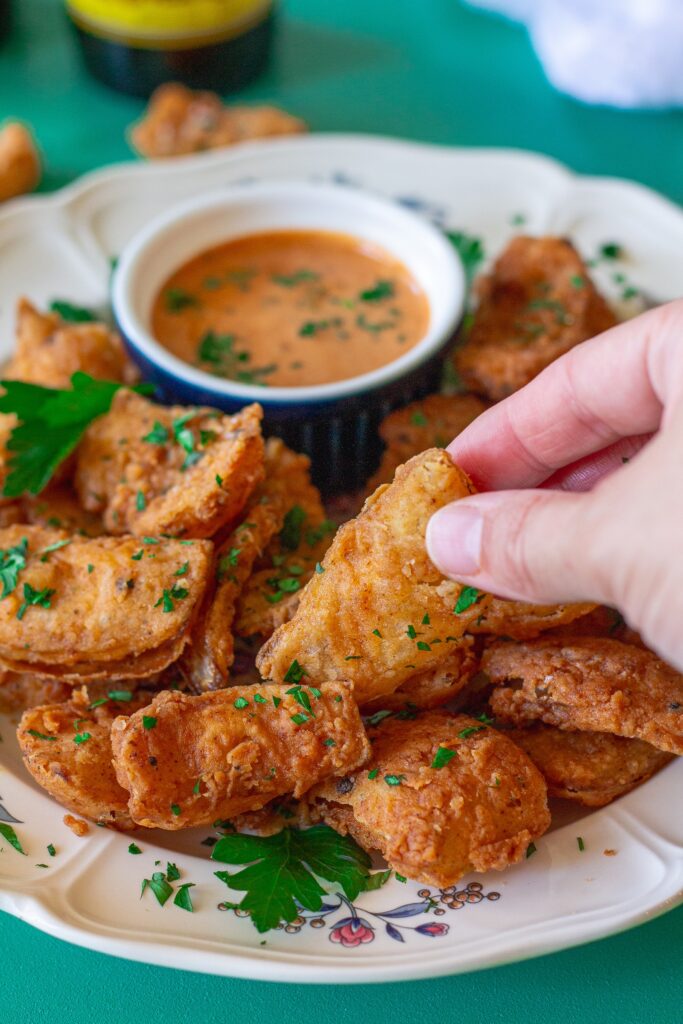 Hand holding a crispy vegan bloomin' onion petal garnished with parsley, ready to be dipped.