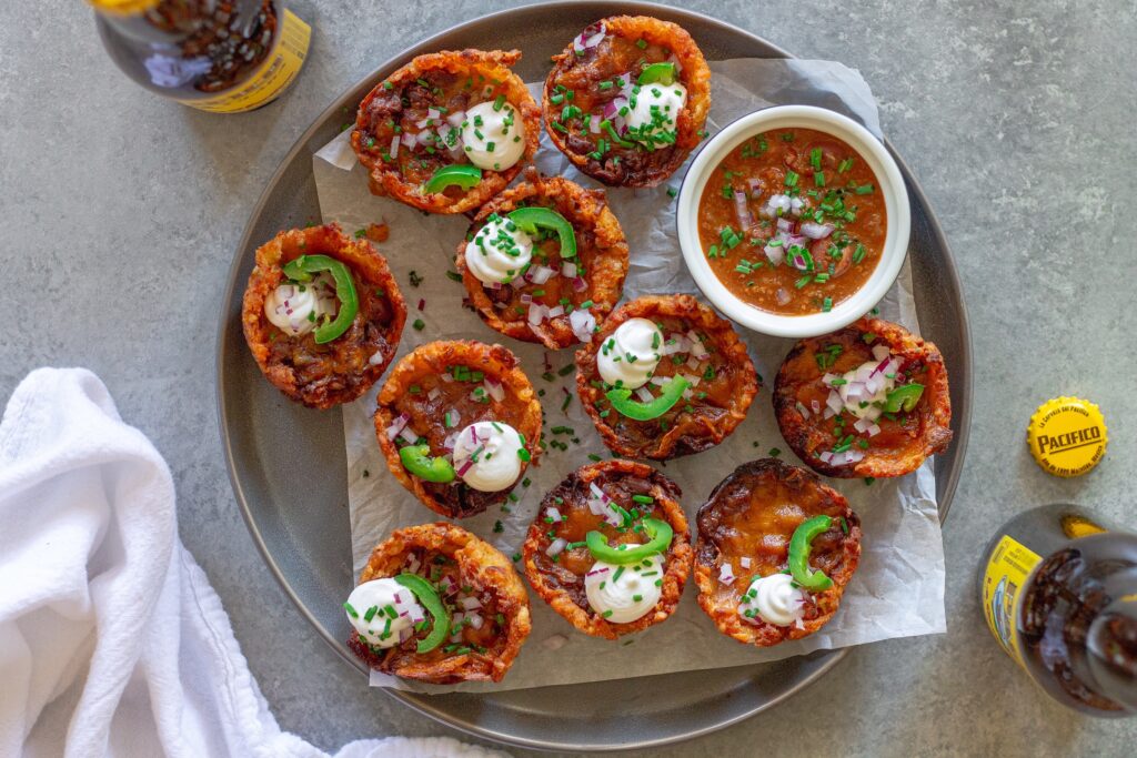 Platter of baked vegan chili cheese tater tot bites, garnished with sliced jalapeños, vegan sour cream, and red onion.