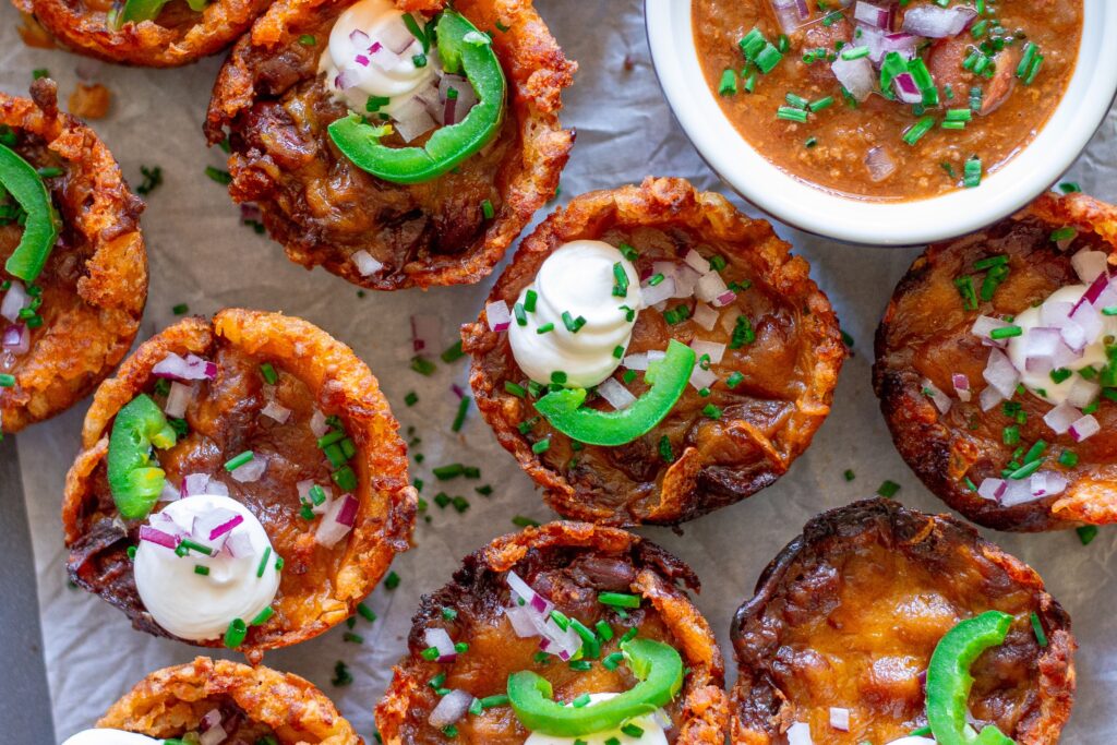 Platter of baked vegan chili cheese tater tot bites, garnished with sliced jalapeños, vegan sour cream, and red onion.