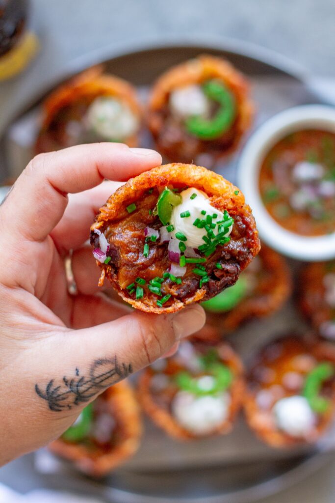 Close-up of a bite-sized vegan chili cheese tater tot cup, topped with vegan sour cream and fresh chives.