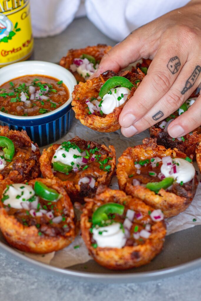 Platter of baked vegan chili cheese tater tot bites, garnished with sliced jalapeños, vegan sour cream, and red onion.