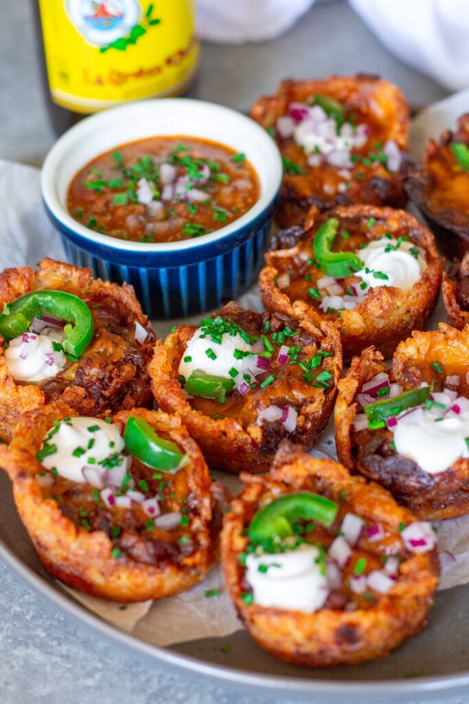 Platter of baked vegan chili cheese tater tot bites, garnished with sliced jalapeños, vegan sour cream, and red onion.