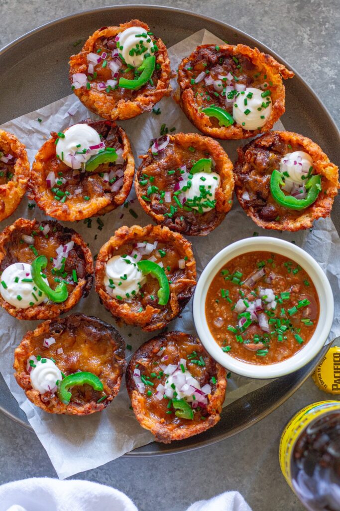 Platter of baked vegan chili cheese tater tot bites, garnished with sliced jalapeños, vegan sour cream, and red onion.