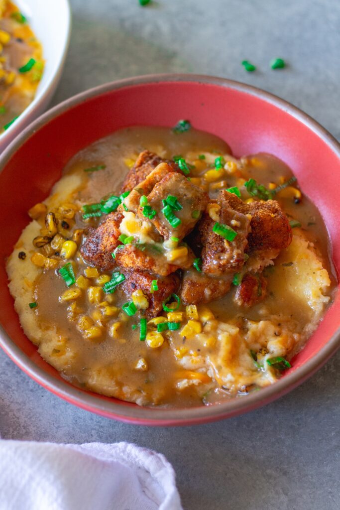 Vegan KFC Copycat Famous Bowl with mashed potatoes, roasted corn, crispy tofu, melted vegan cheese, and gravy in a bowl, garnished with green onions.