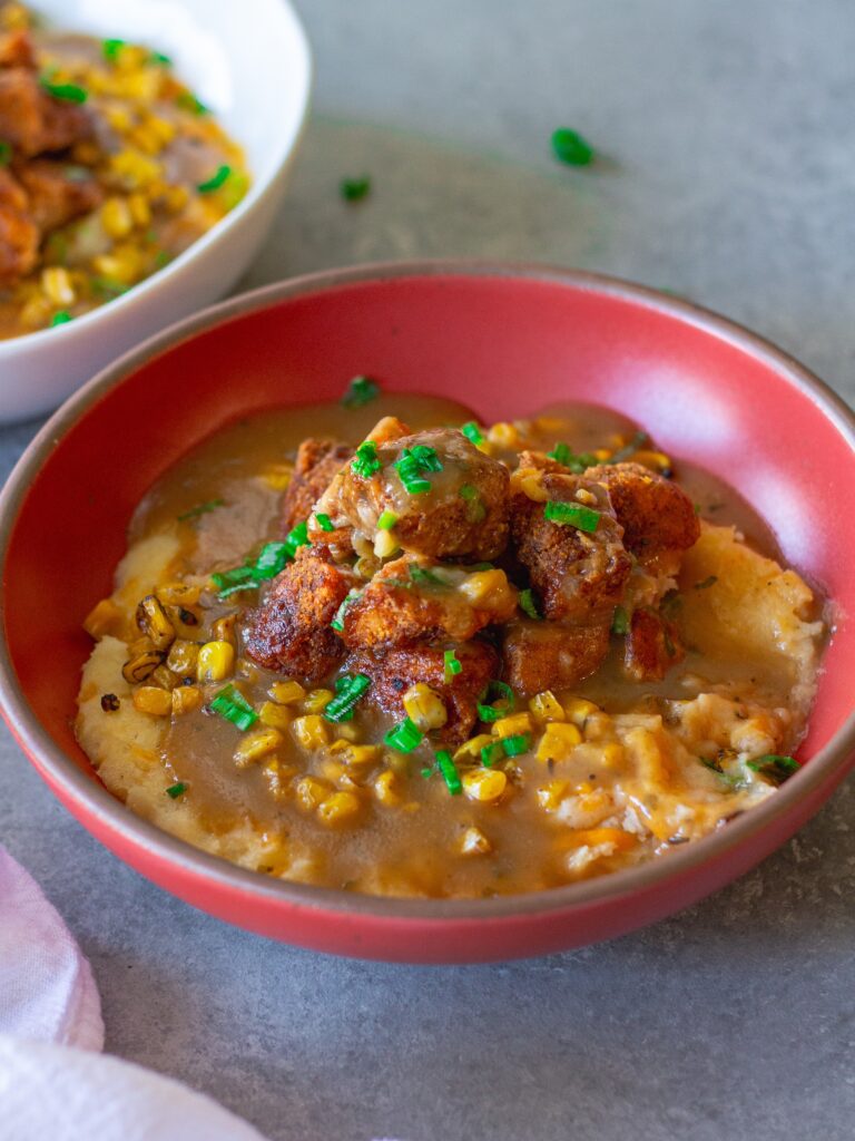 Vegan KFC Copycat Famous Bowl with mashed potatoes, roasted corn, crispy tofu, melted vegan cheese, and gravy in a bowl, garnished with green onions.