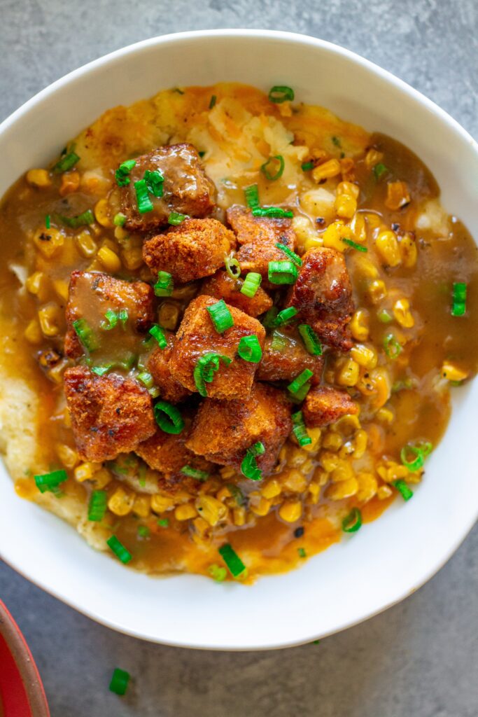 Vegan KFC Copycat Famous Bowl with mashed potatoes, roasted corn, crispy tofu, melted vegan cheese, and gravy in a bowl, garnished with green onions.