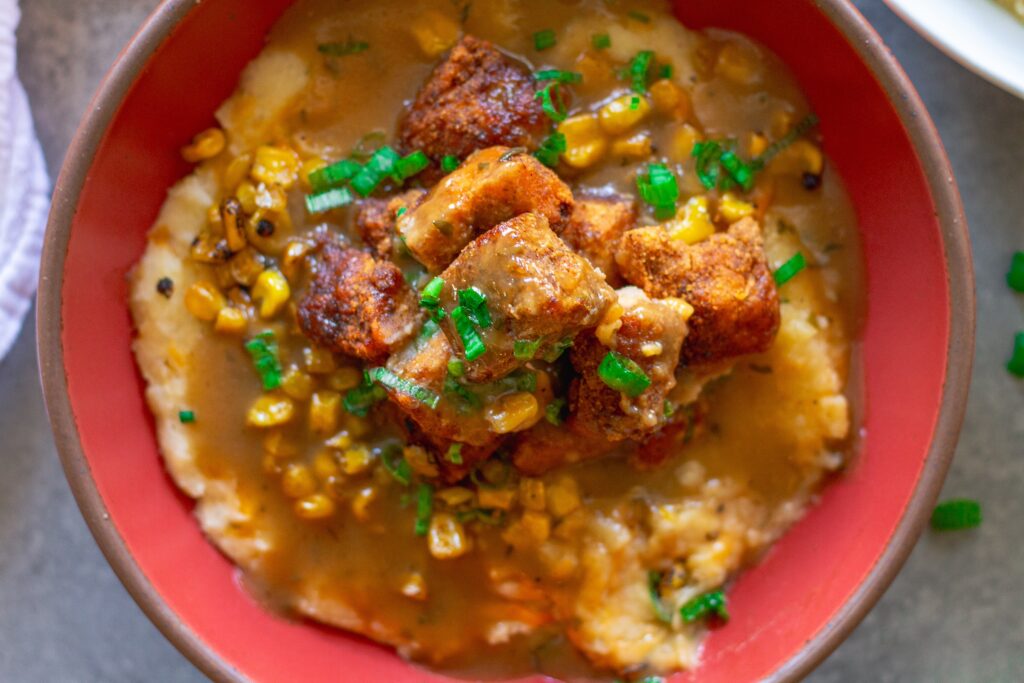 Vegan KFC Copycat Famous Bowl with mashed potatoes, roasted corn, crispy tofu, melted vegan cheese, and gravy in a bowl, garnished with green onions.