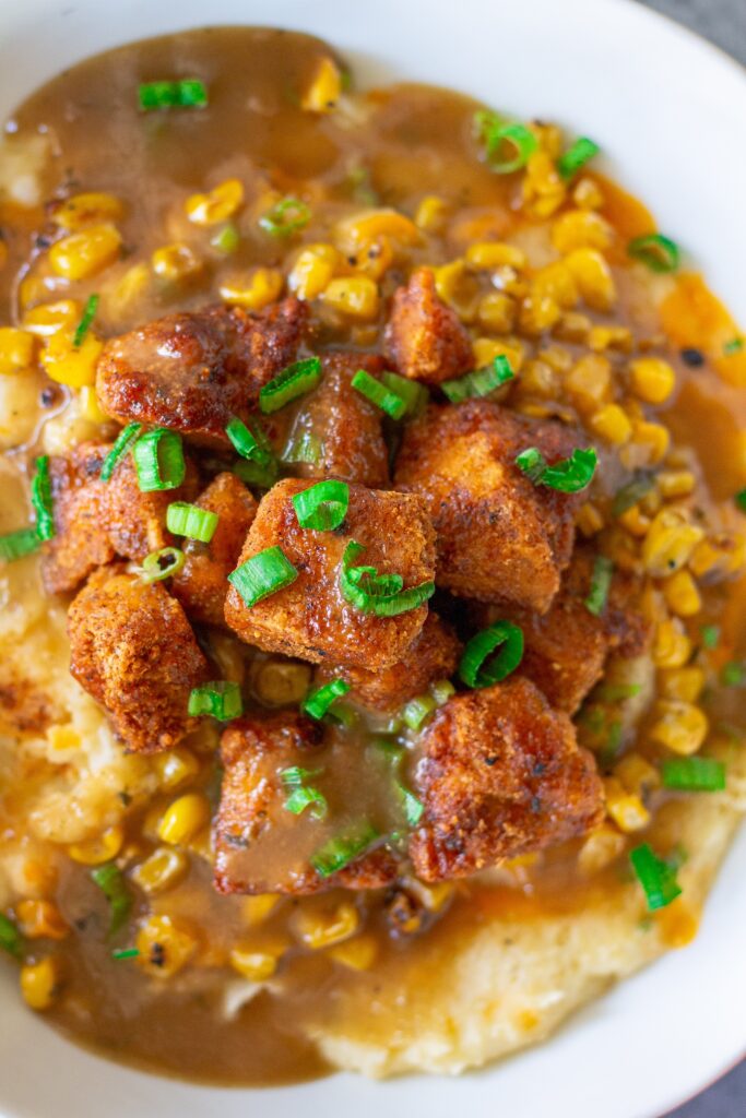 Vegan KFC Copycat Famous Bowl with mashed potatoes, roasted corn, crispy tofu, melted vegan cheese, and gravy in a bowl, garnished with green onions.