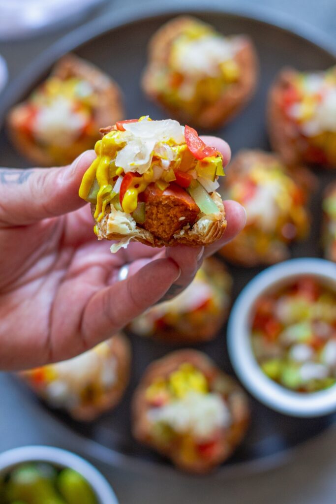 A hand holding a mini vegan Chicago hot dog bite topped with classic Chicago-style toppings like tomatoes and mustard.