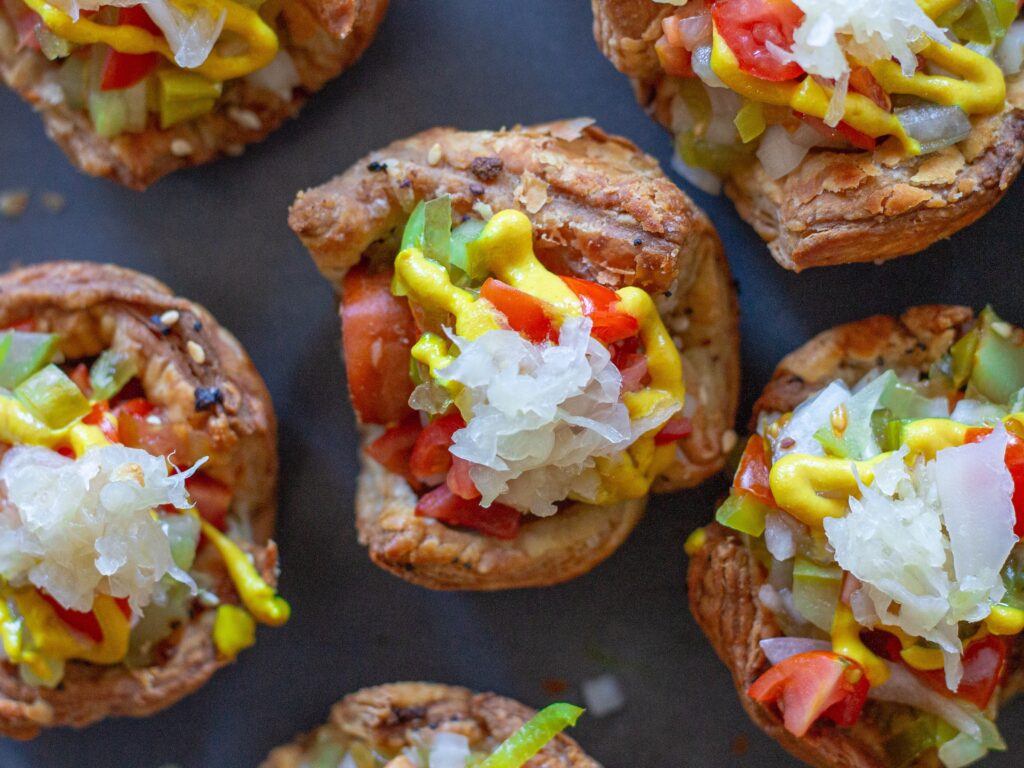 Close-up of a single mini vegan Chicago hot dog bite topped with diced tomatoes, pickles, and a drizzle of mustard