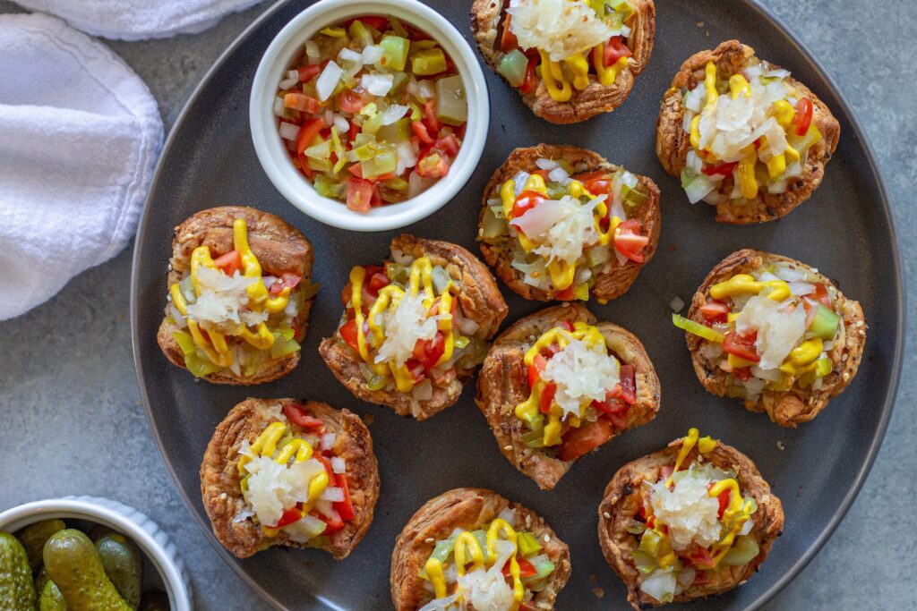 Mini vegan Chicago hot dog bites with toppings like diced tomato, pickles, and mustard, served on a platter.