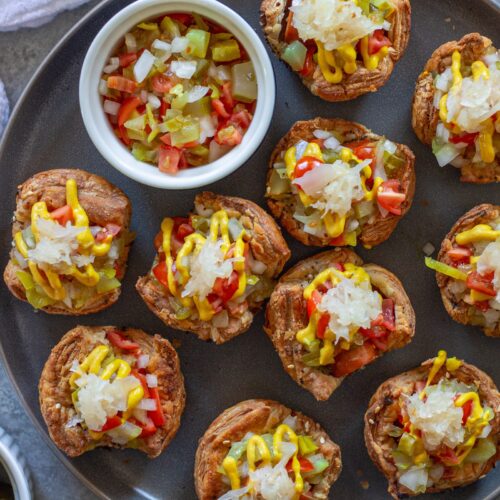 Mini vegan Chicago hot dog bites with toppings like diced tomato, pickles, and mustard, served on a platter.