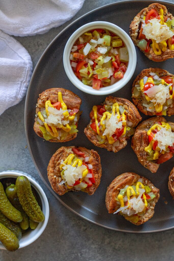 Mini vegan Chicago hot dog bites with toppings like diced tomato, pickles, and mustard, served on a platter.