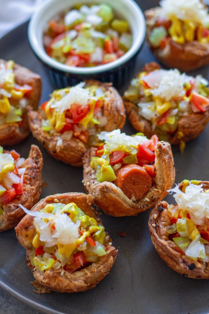 Mini vegan Chicago hot dog bites with toppings like diced tomato, pickles, and mustard, served on a platter.