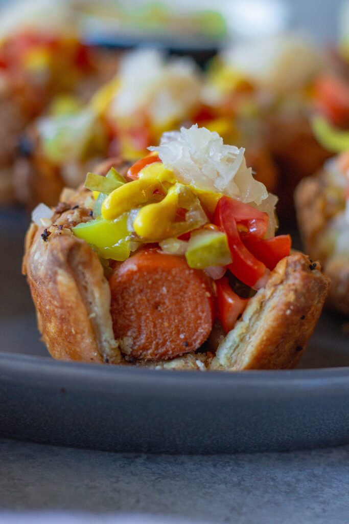 Close-up of a single mini vegan Chicago hot dog bite topped with diced tomatoes, pickles, and a drizzle of mustard.